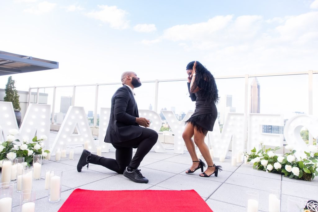 A stunning rooftop proposal at Ventanas, Atlanta, GA, with a man on one knee and "Marry Me" marquee letters.